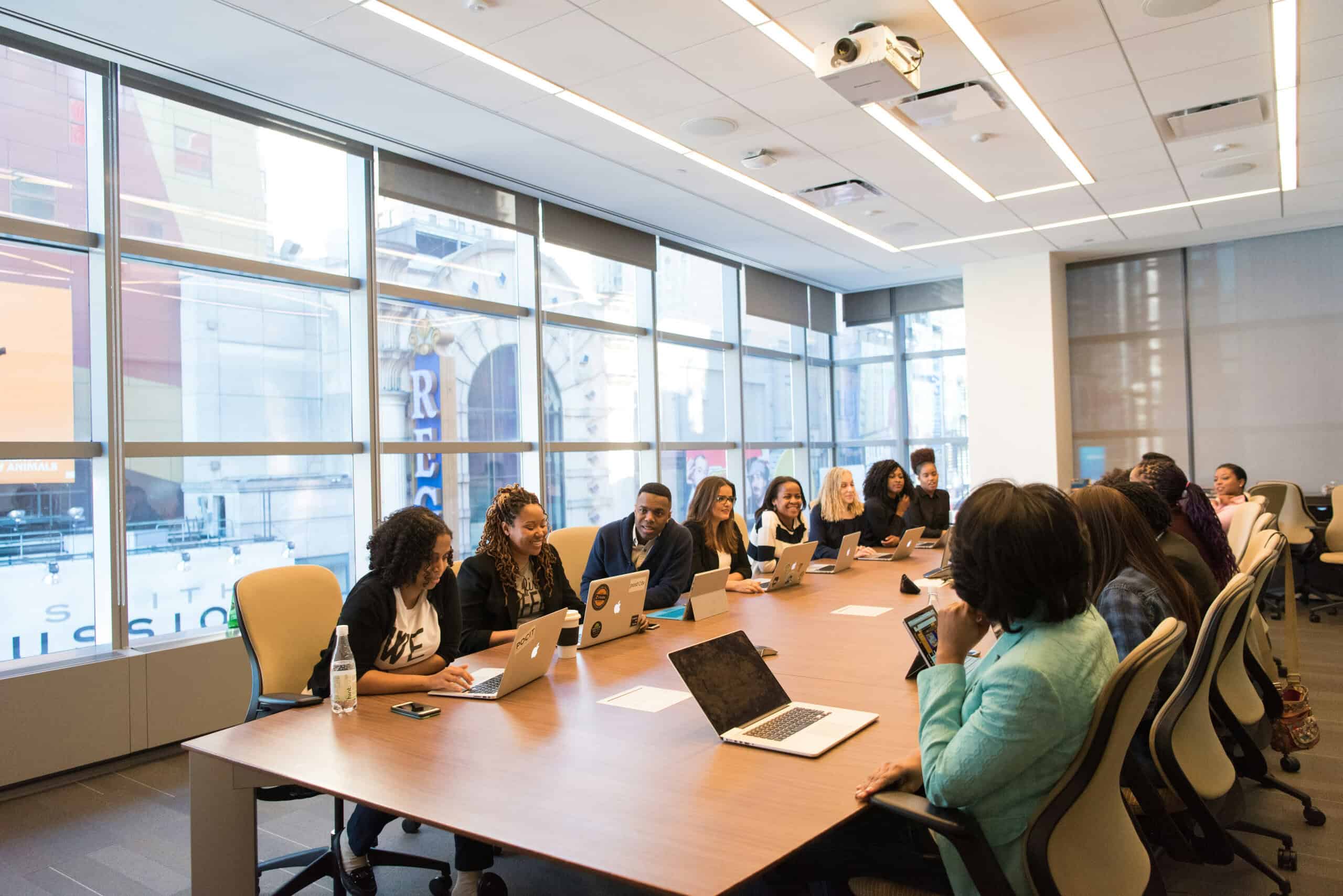 A diverse group of people sit around a large conference table with laptops during a meeting in a bright, modern office.