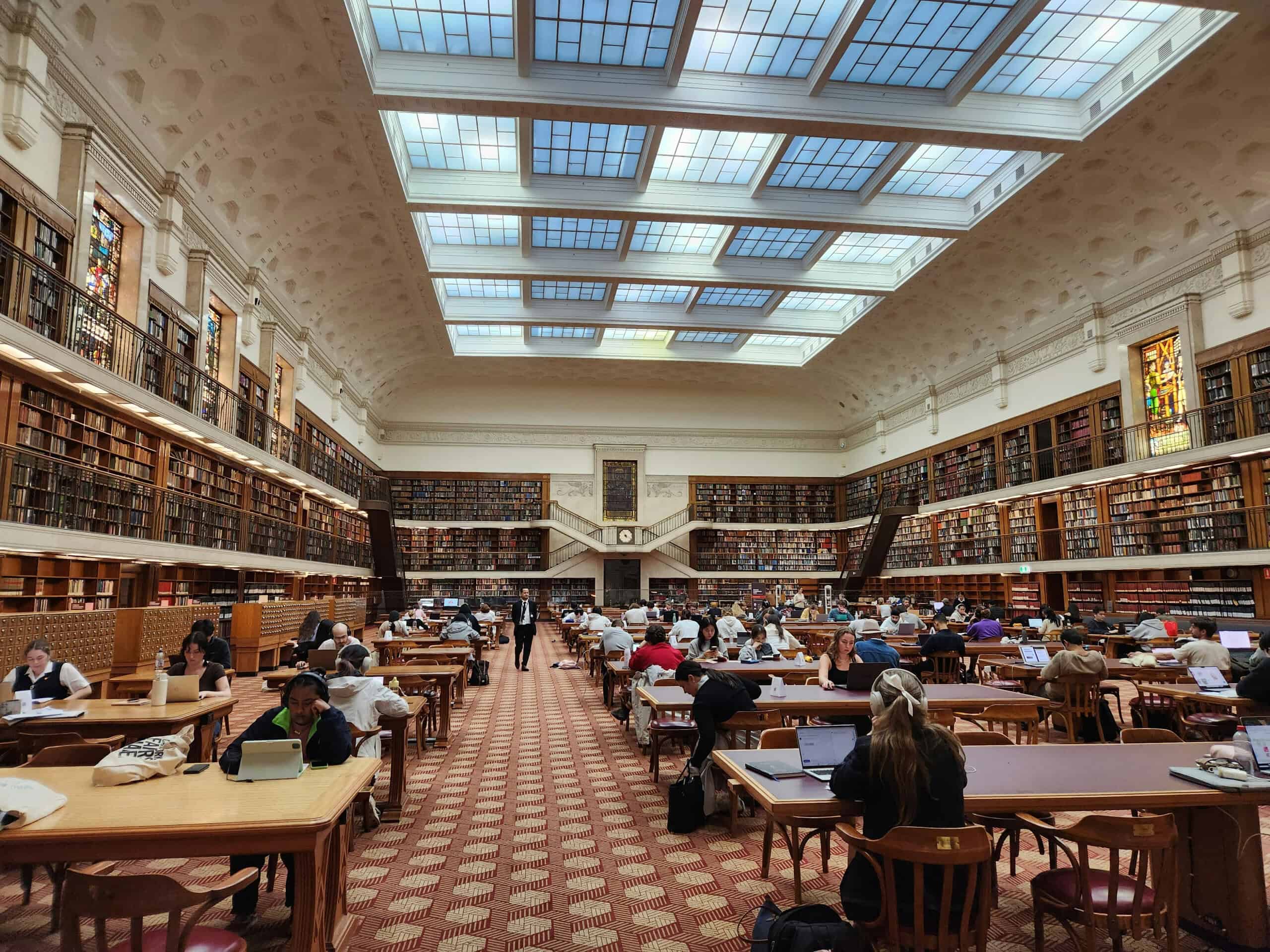 People study and work on laptops in a grand library reading room with high ceilings, stained glass, and walls lined with bookshelves.